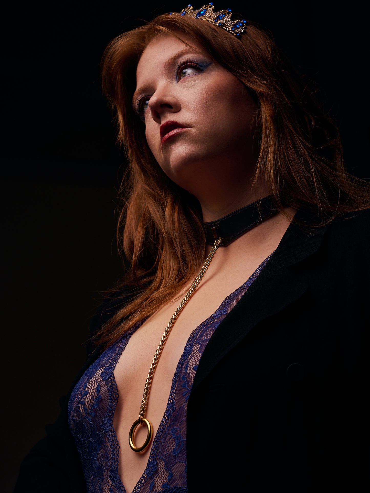 Woman wearing a blue lace top and black blazer with a black leather necklace with gold chain against a dark background