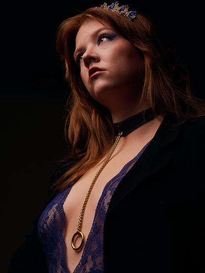 Woman wearing a blue lace top and black blazer with a black leather necklace with gold chain against a dark background