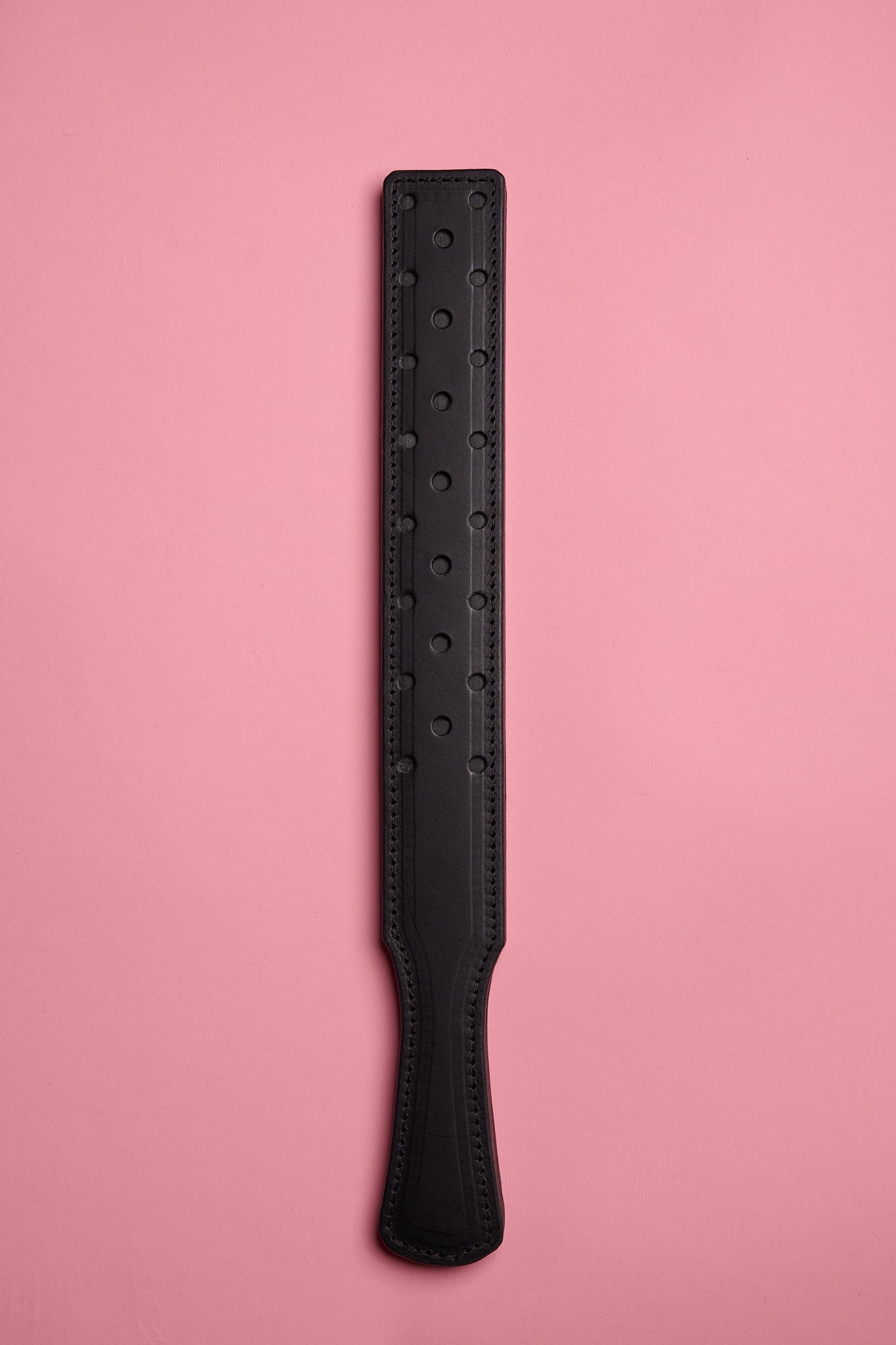 A black leather paddle with a fenestrated surface, laid on a pink background.