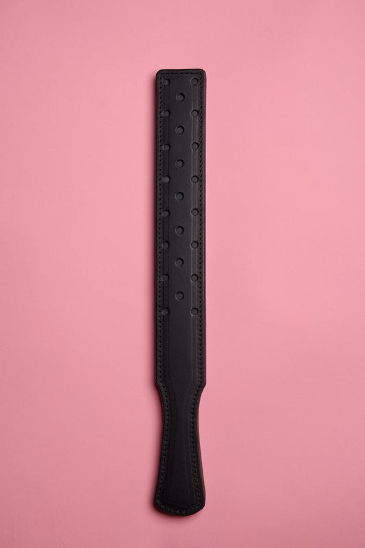 A black leather paddle with a fenestrated surface, laid on a pink background.