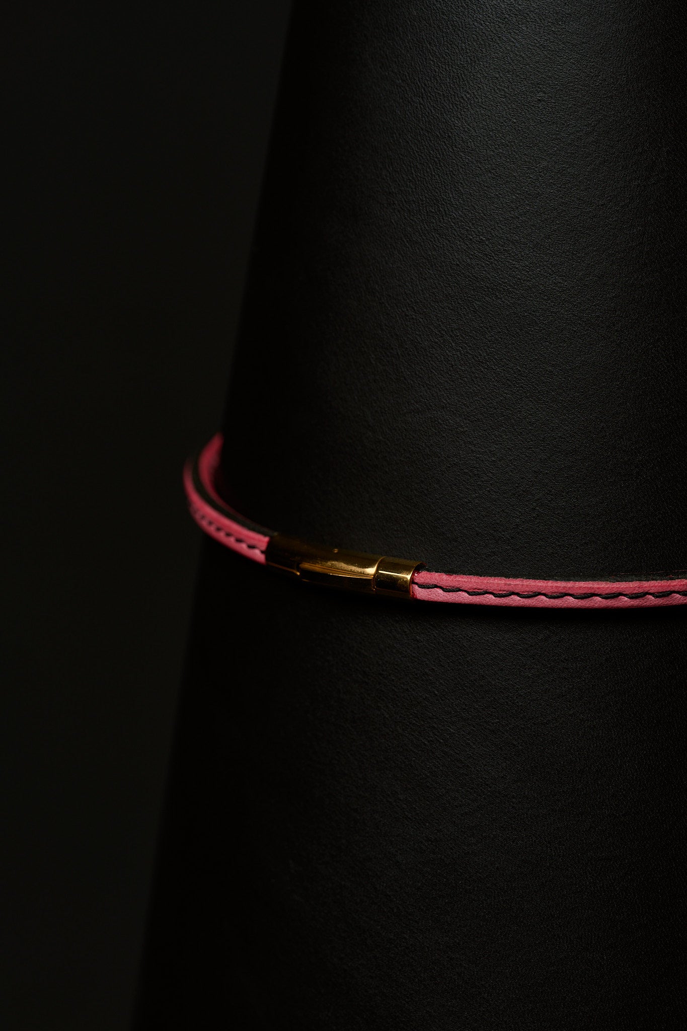Thin pink necklace with black stitching and a gold clasp close up on a black background.