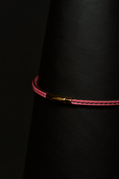 Thin pink necklace with black stitching and a gold clasp close up on a black background.